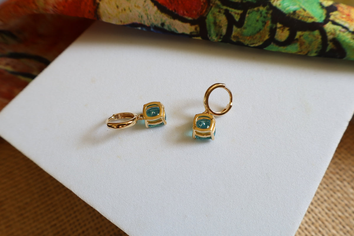 Custom 18K yellow gold huggie hoop earrings featuring cushion-cut ocean-blue gemstones in a dangling drop design, inspired by the beauty of the sea.