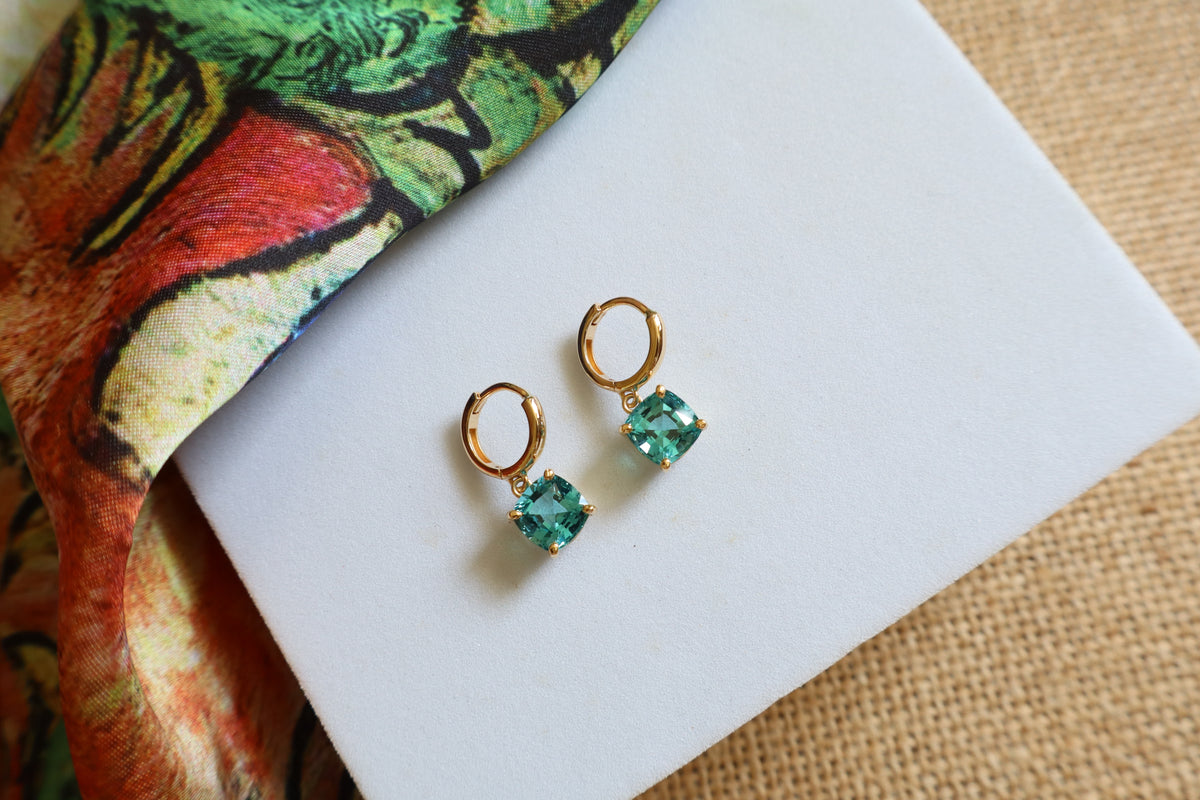 Custom 18K yellow gold huggie hoop earrings featuring cushion-cut ocean-blue gemstones in a dangling drop design, inspired by the beauty of the sea.