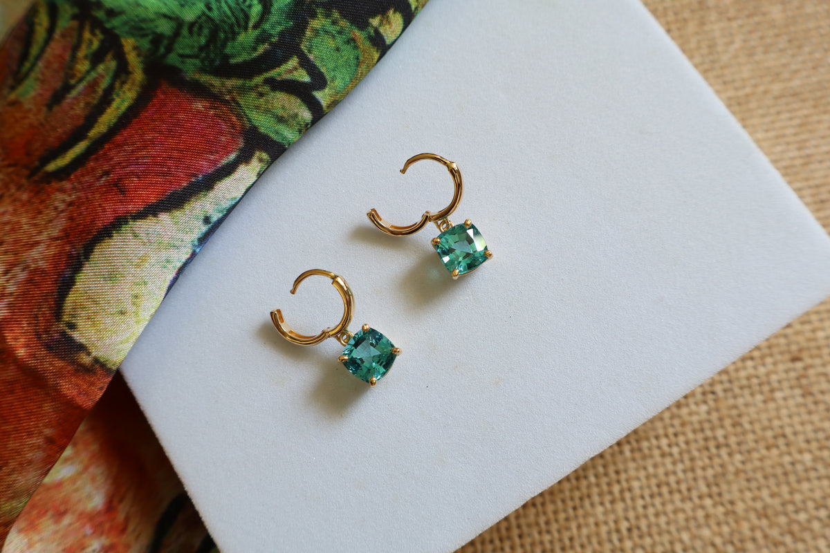Custom 18K yellow gold huggie hoop earrings featuring cushion-cut ocean-blue gemstones in a dangling drop design, inspired by the beauty of the sea.