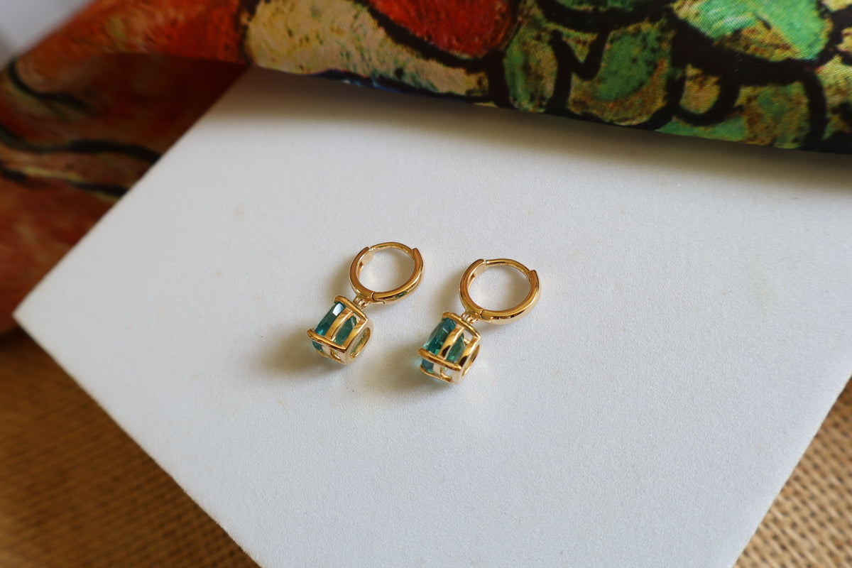Custom 18K yellow gold huggie hoop earrings featuring cushion-cut ocean-blue gemstones in a dangling drop design, inspired by the beauty of the sea.
