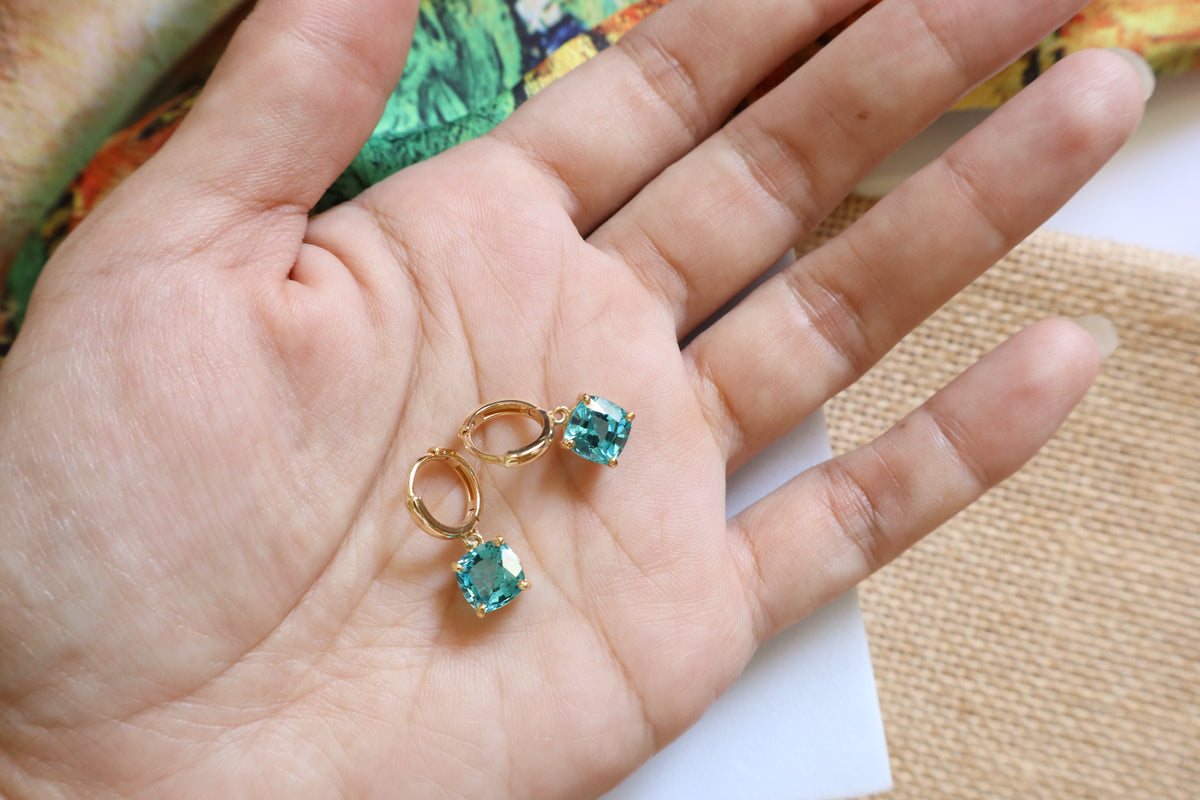 Custom 18K yellow gold huggie hoop earrings featuring cushion-cut ocean-blue gemstones in a dangling drop design, inspired by the beauty of the sea.