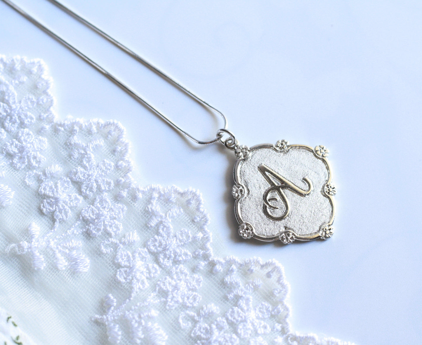 A white gold necklace with a diamond-shaped pendant. With an "A" initial with flower borders in it.