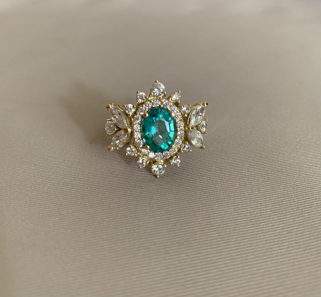 An oval paraiba tourmaline with marquise and round stones around it. Set in yellow gold.