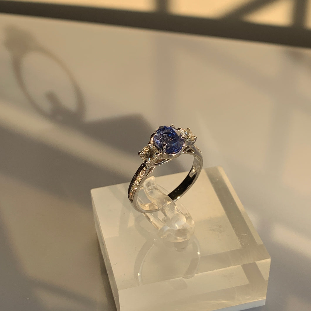 An oval blue sapphire with two round diamonds on its sides. All uplifted with 3 intersected prongs. Combined with small brilliant diamonds as its shoulders. All set in white gold.